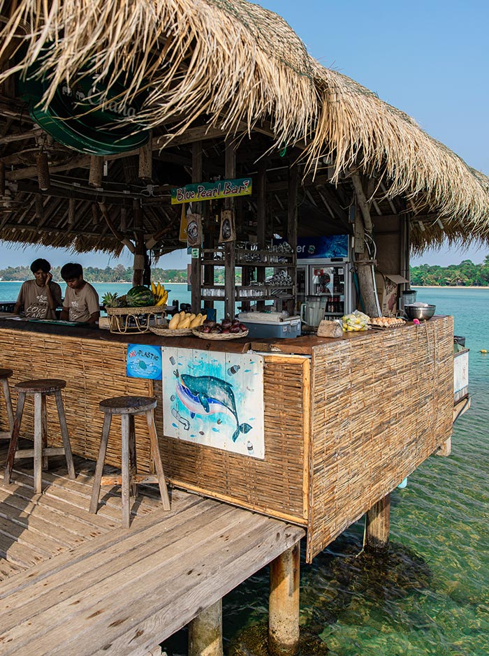 Thatched roof beach bar on wooden stilts over clear water with a bamboo counter and stools along the edge. Two staff stand behind the bar beneath a sign reading "Blue Pearl Bar" with fresh fruit and drinks displayed.