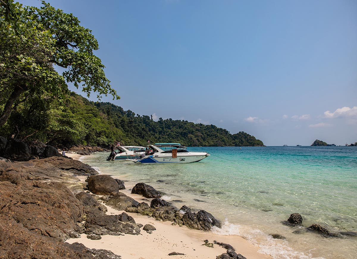 Rocky shoreline with dark stones and soft sand meets clear turquoise water where two small motorboats are anchored near the beach. Lush green trees line the coast and a forested island rises in the background under a bright blue sky, creating a calm tropical seaside scene.