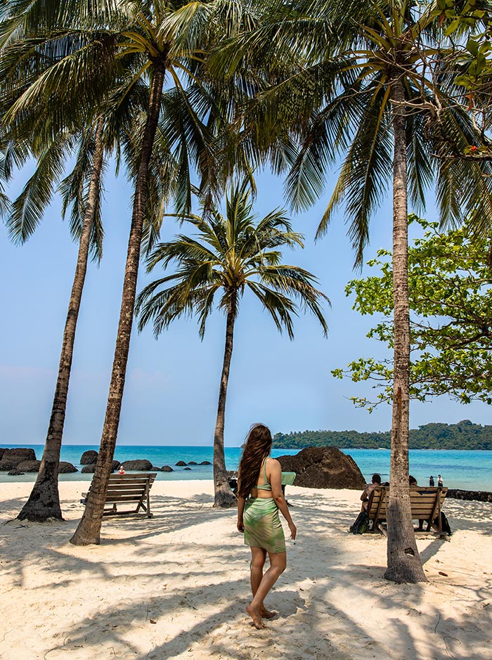 Woman in a light green dress walks barefoot across white sand toward the ocean, framed by tall palm trees and wooden benches with a few people sitting in the shade. Turquoise water, scattered rocks, and a distant green island sit under a clear blue sky, creating a relaxed tropical beach scene.