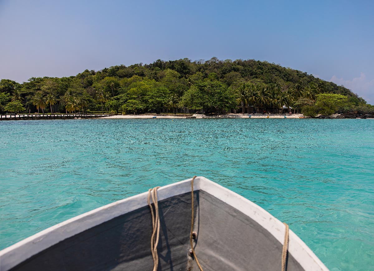 View from the front of a small boat approaching a lush green island with palm trees along the shoreline. Clear turquoise water surrounds the island under a bright blue sky.