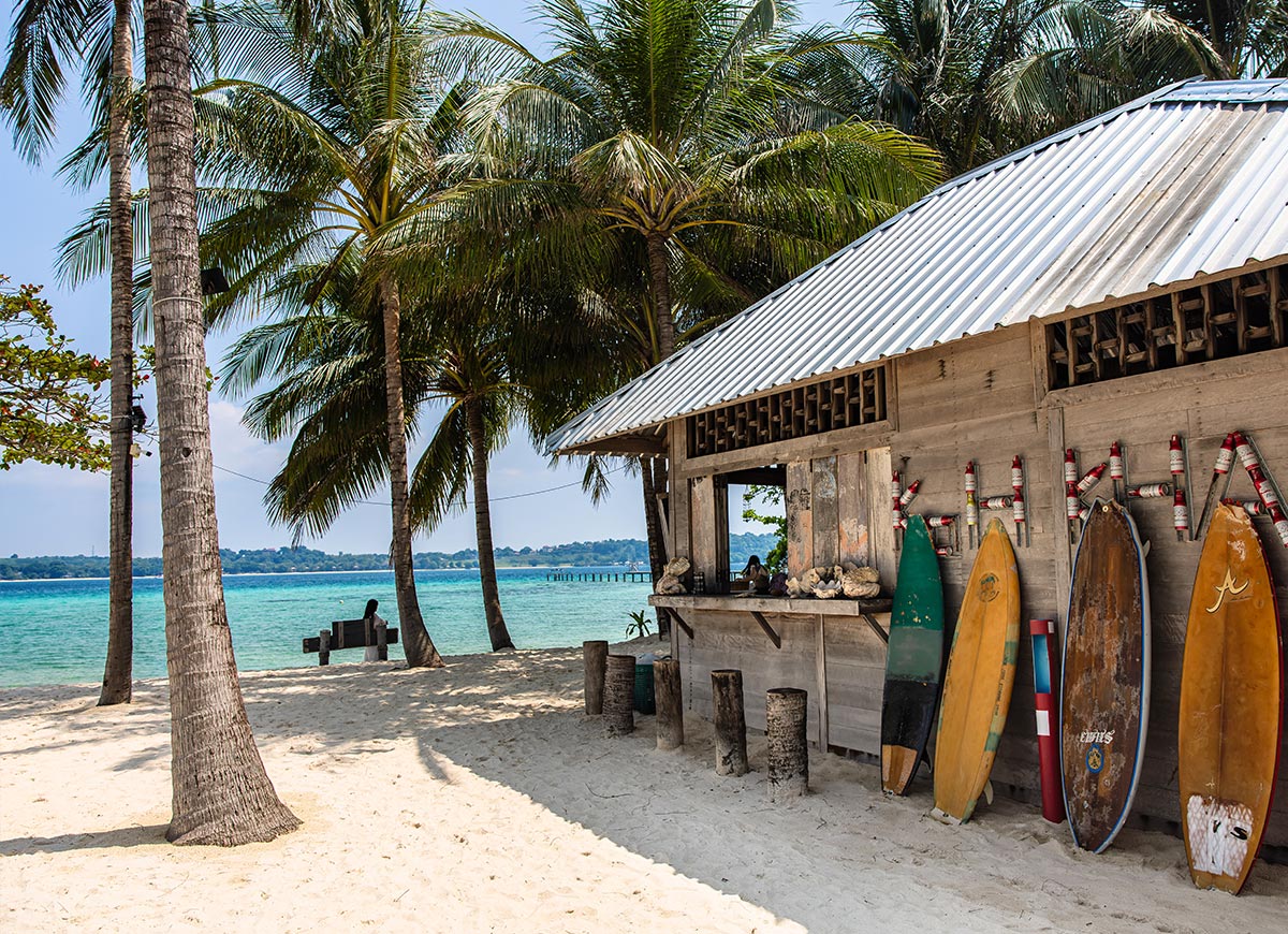 Beachside wooden hut with a corrugated metal roof and a row of surfboards leaning against the wall on sandy ground. Tall palm trees frame the scene with clear turquoise water and a distant pier visible in the background.