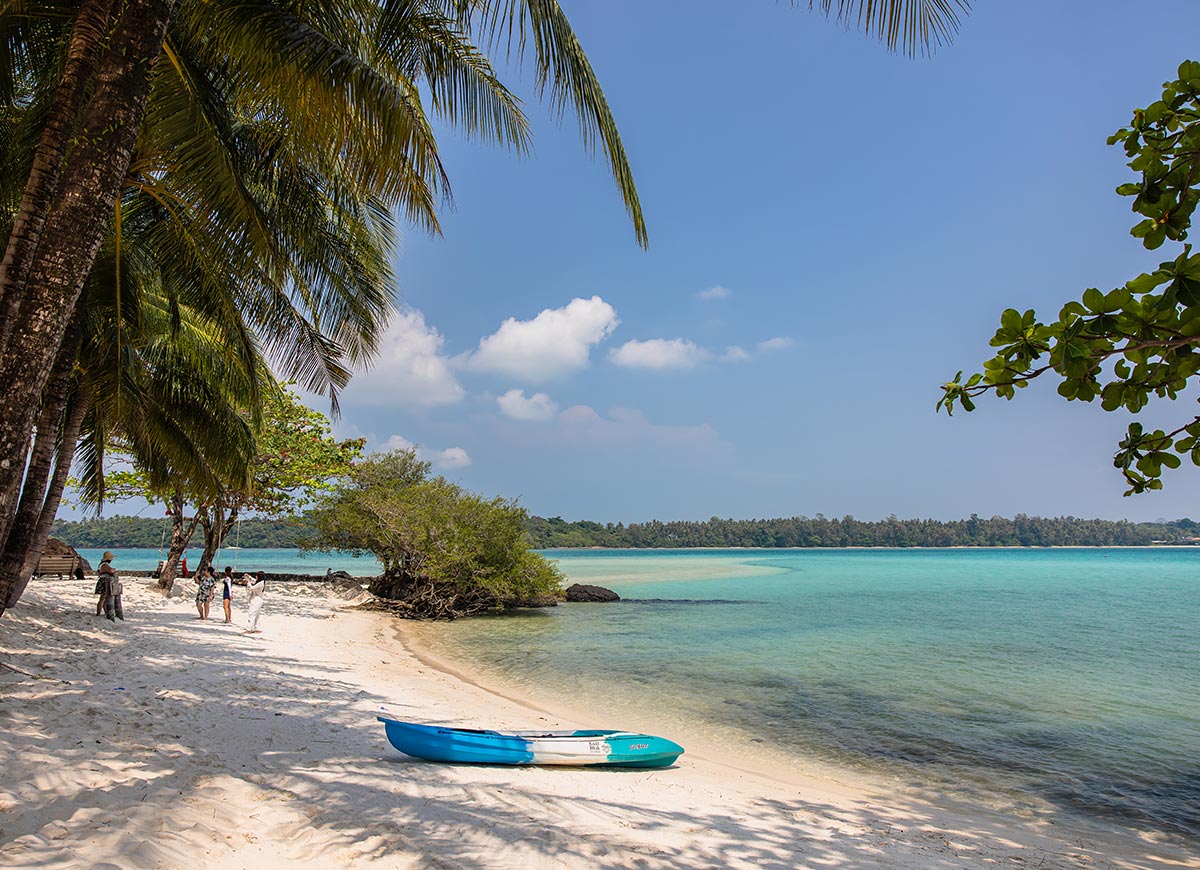 White sand beach lined with palm trees and clear turquoise water gently meeting the shore. A blue kayak rests on the sand while a few people walk along the coastline under a bright blue sky.