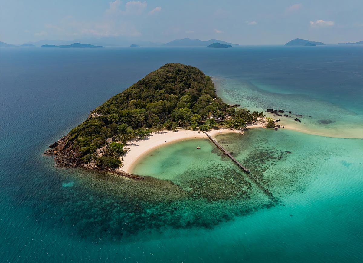 Aerial view of a small tropical island with white sand beaches and lush green trees surrounded by clear turquoise water. A long wooden pier extends into the sea while distant islands fade into the horizon.