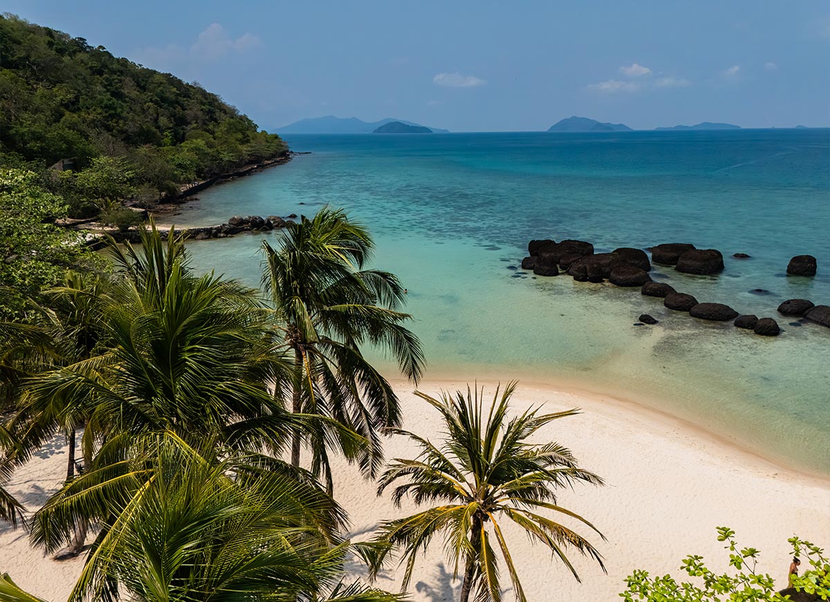 Secluded tropical beach with soft white sand and tall palm trees overlooking clear turquoise water. Dark rocks sit just offshore while forested hills and distant islands stretch across the horizon under a bright sky.