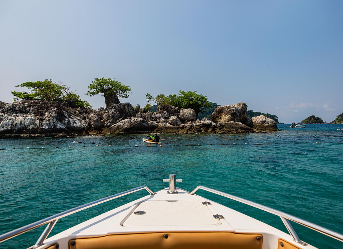View from the front of a boat looking out over clear turquoise water toward a rocky island with scattered trees. A few people snorkel near the rocks while another boat floats in the distance under a bright blue sky.