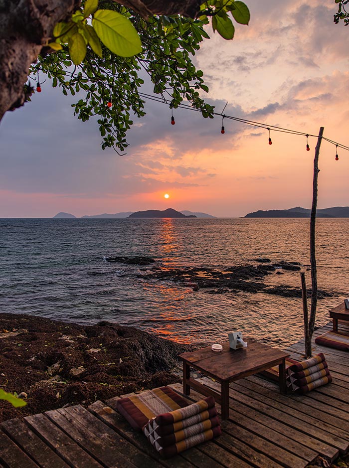 Wooden seaside deck with low table and floor cushions overlooking rocky shoreline at sunset. The sun dips toward the horizon casting orange reflections on the water while string lights and leafy branches frame distant island silhouettes.