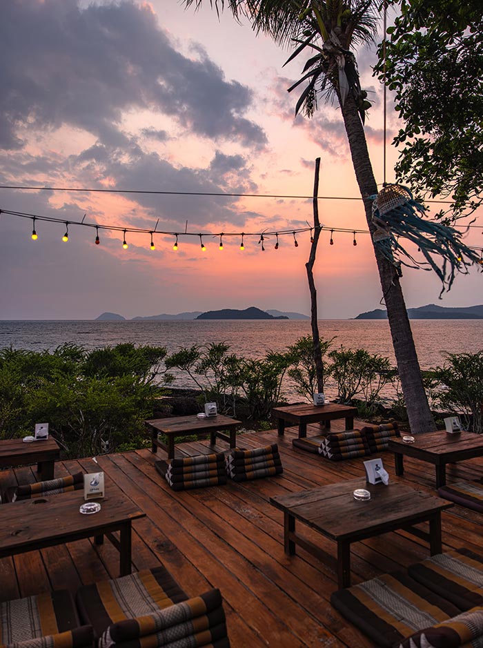 Wooden deck seating by the ocean at sunset with low tables and floor cushions arranged under string lights. A palm tree leans overhead while the sky glows pink and orange above calm water and distant islands.
