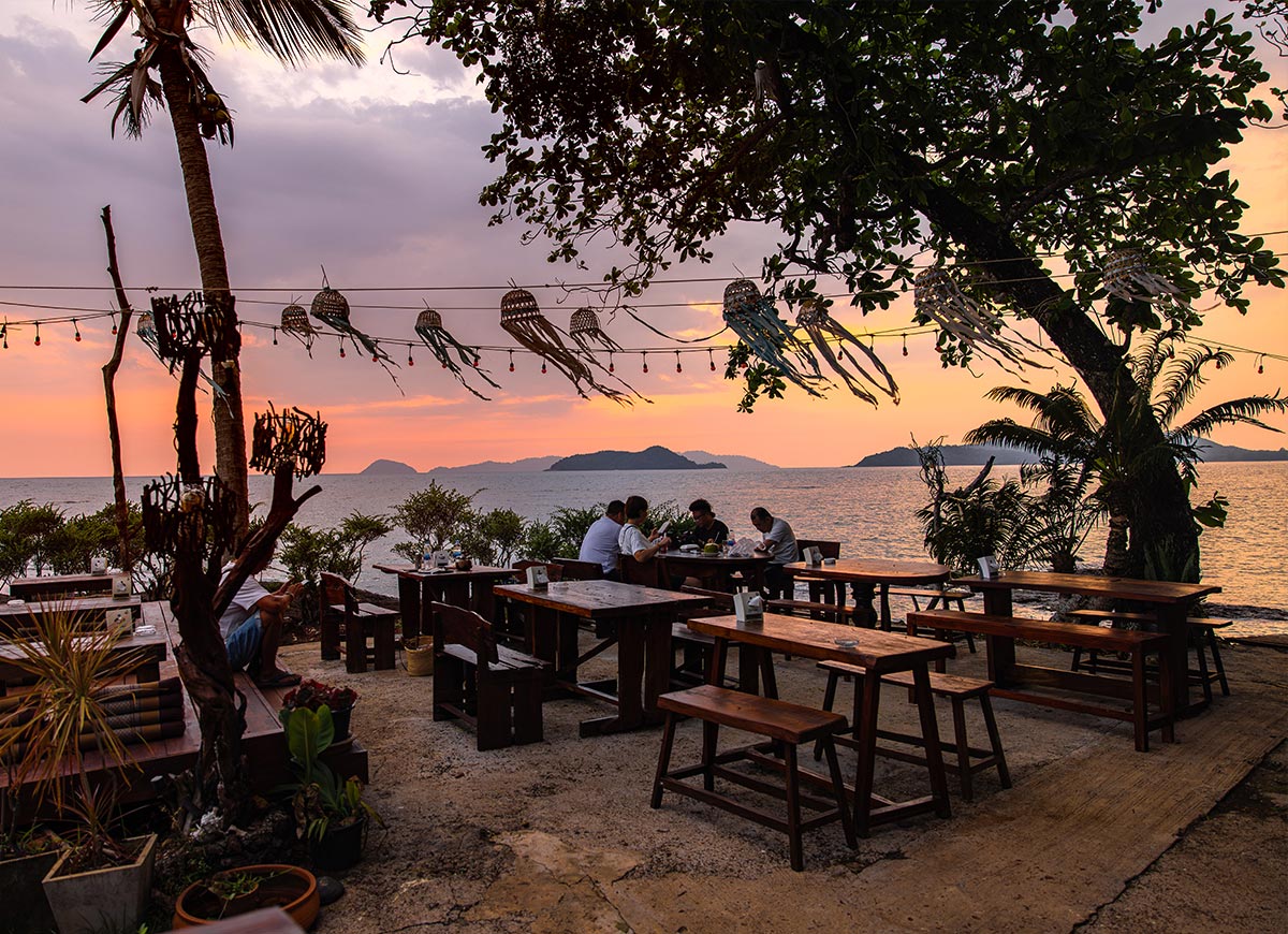 Beachfront restaurant at sunset with wooden tables and benches set on sand beneath trees, string lights and hanging lanterns swaying overhead. A small group of people sits eating by the water as the sky glows orange and pink over distant islands.