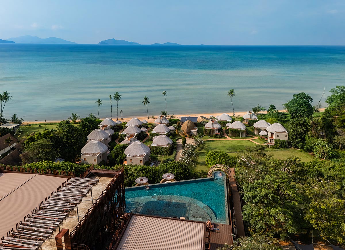 Elevated view of a beachfront resort with thatched villas surrounded by greenery and palm trees, overlooking calm turquoise water and distant islands. An infinity pool in the foreground blends into the ocean horizon, highlighting the coastal setting.