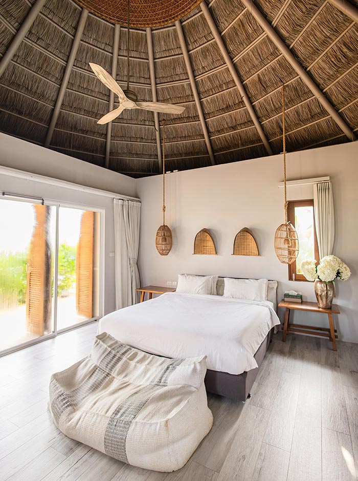 Bright bedroom with a thatched dome ceiling and wooden beams, featuring a white bed, woven pendant lights, and a large sliding glass door opening to greenery. A soft beanbag chair and simple wooden furniture add to the airy tropical design.