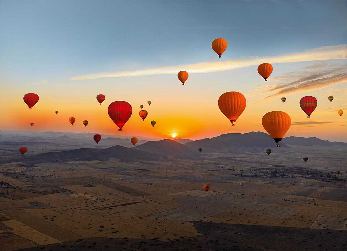 Dozens of colorful hot air balloons float above a desert landscape at sunrise, with the sun rising behind distant mountains and casting a warm golden glow across the sky and valleys below.