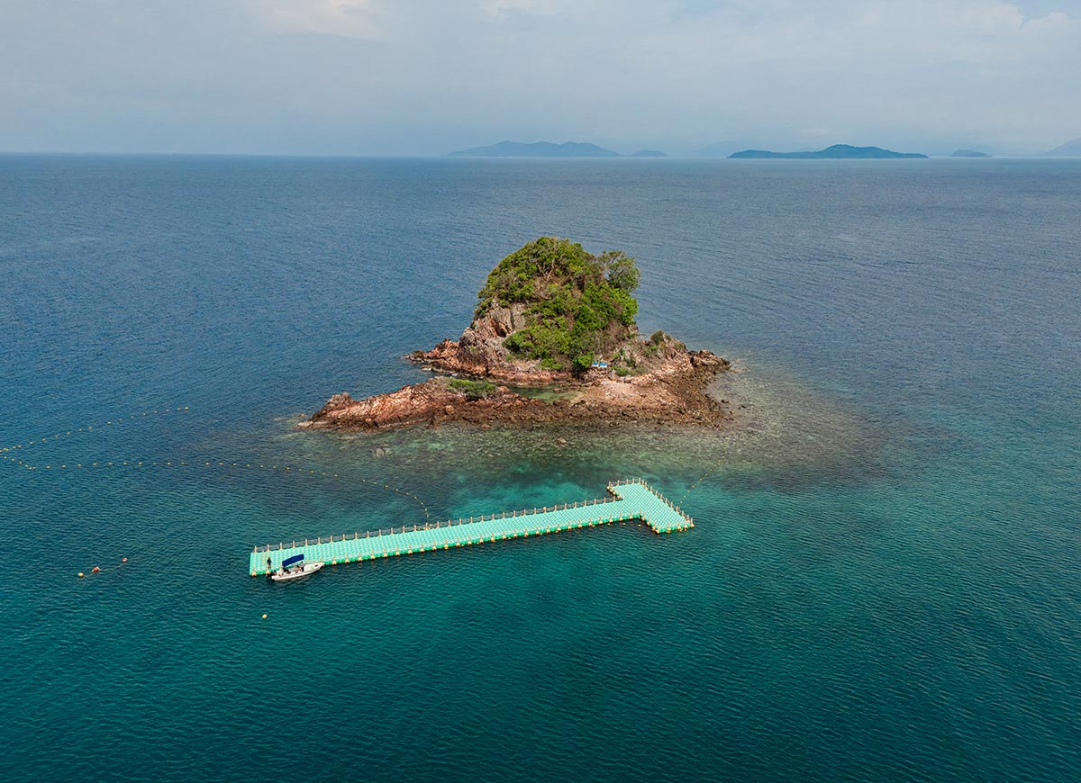 Aerial view of a small rocky island covered in greenery surrounded by deep blue ocean. A floating pier extends toward the island with a small boat docked at the end while distant islands fade into the horizon.