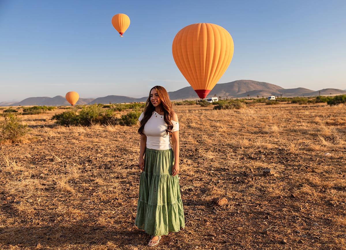 A woman stands in a sunlit desert as hot air balloons lift off around her, with golden morning light illuminating the landscape and mountains in the background.