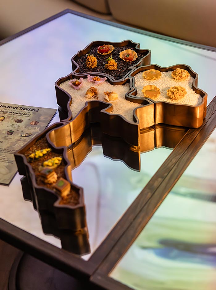 Map shaped serving tray presenting small plated bites arranged across sections filled with rice and grains, designed to resemble Thailand. A card beside it reads “Taste of Thailand” and “Regional Tasting Bites,” adding context to the curated culinary experience. The artistic presentation highlights regional flavors in a visually engaging and organized layout.