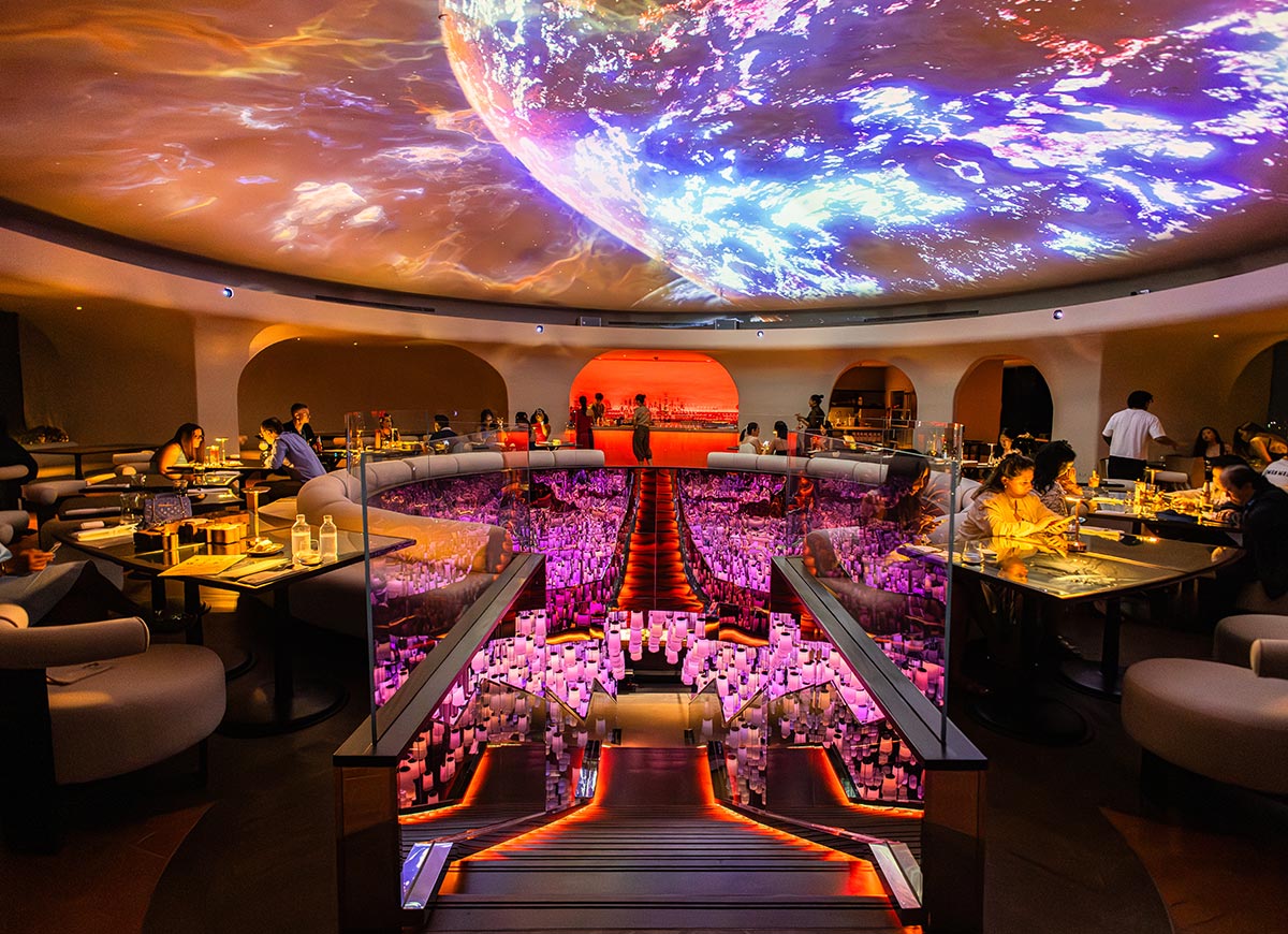 Futuristic restaurant interior with diners seated around curved tables overlooking a glowing central installation of suspended lights. A dramatic ceiling projection of a colorful planet spans overhead, casting vibrant hues across the space. Illuminated stairs lead down through the centerpiece, creating a striking, immersive dining atmosphere.