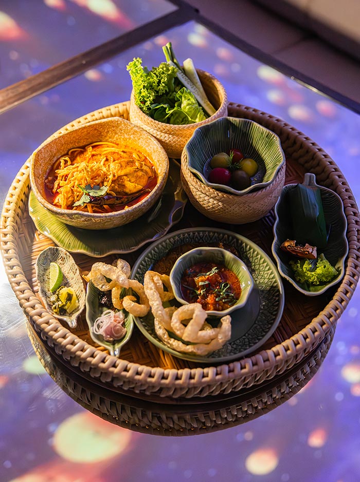 Woven tray filled with a variety of plated dishes including curry noodles, fresh vegetables, dips, and crispy snacks arranged in small bowls. Colorful ingredients and garnishes create a vibrant presentation, with sauces and herbs adding detail to each dish. The setting feels like a curated tasting experience with multiple small portions to sample.