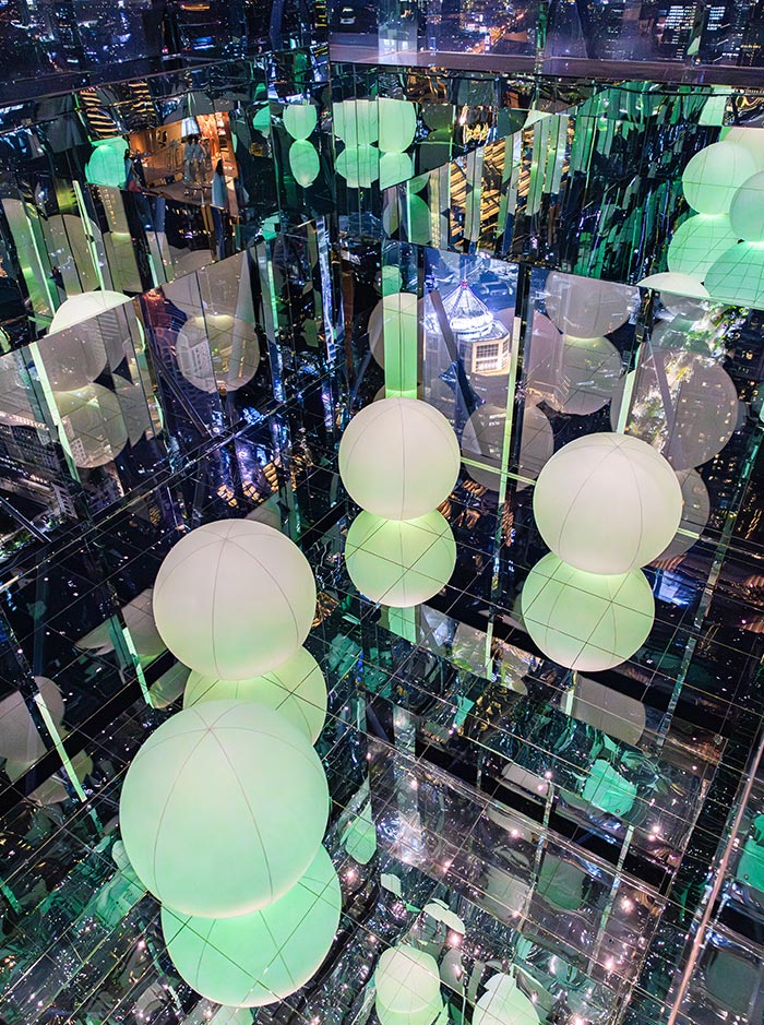 Mirrored room filled with glowing spherical lights in soft green tones, suspended at different heights and reflected endlessly on glass surfaces. The reflections create an illusion of depth, with a city skyline visible through the surrounding windows. The space feels immersive and futuristic, blending light, reflection, and urban views.