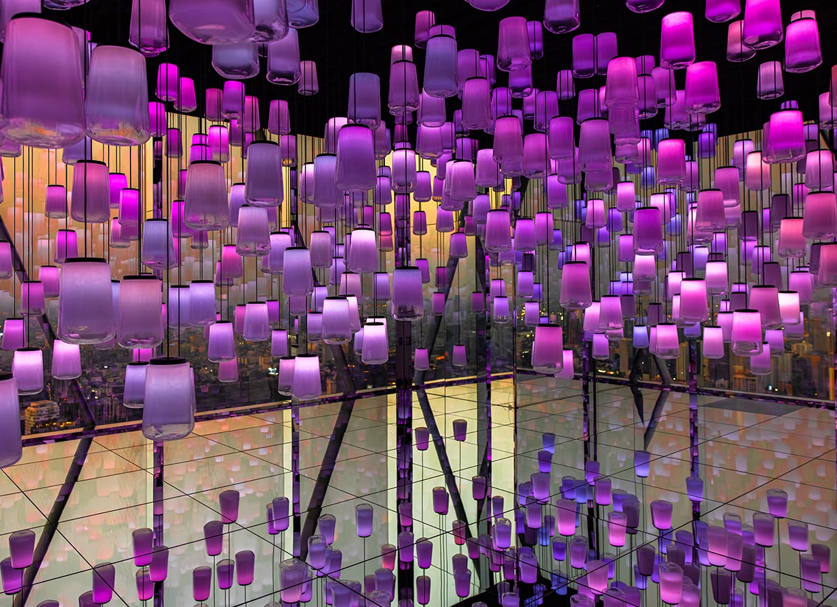 Immersive installation of hanging lantern style lights glowing in shades of purple and pink, suspended at varying heights in a mirrored room. The reflective floor and walls multiply the lights, creating a surreal infinity effect. City views are faintly visible through the glass, adding depth to the vibrant, dreamlike space.