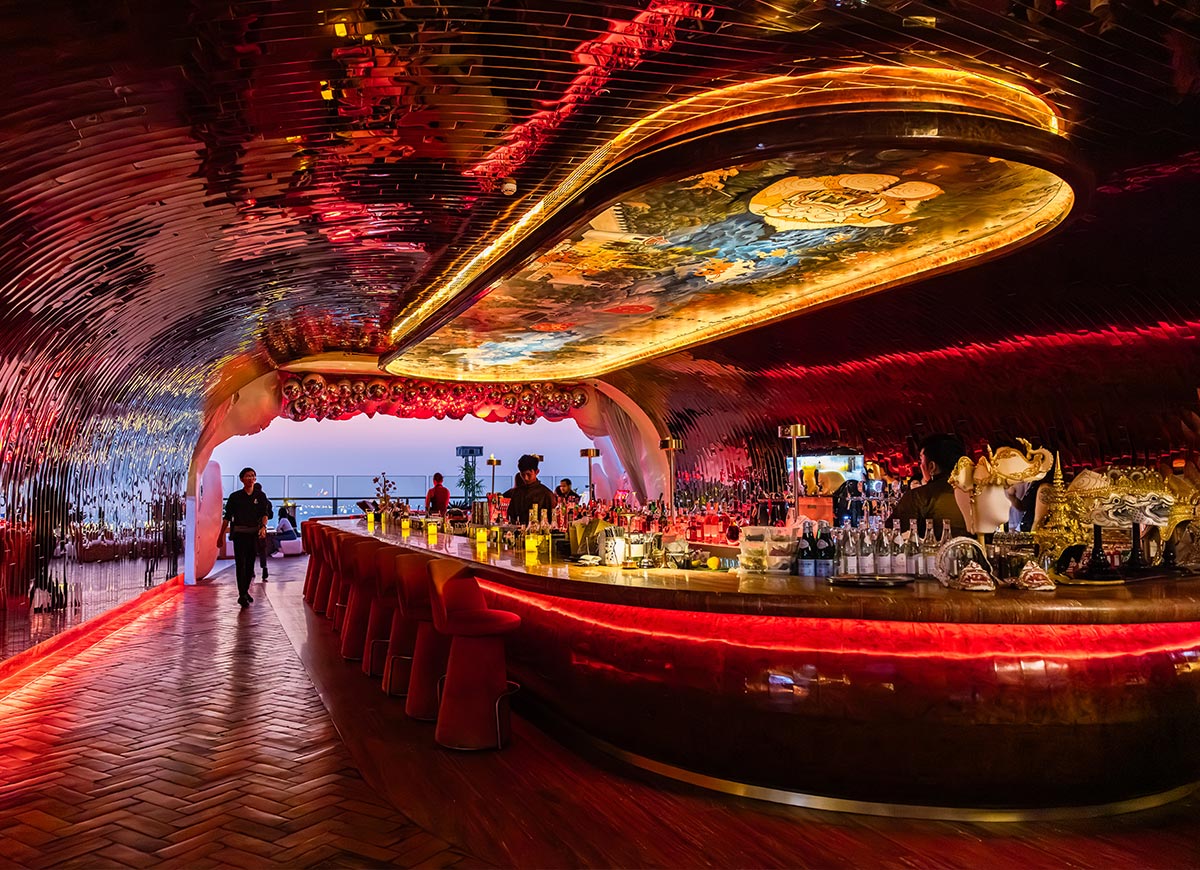 Futuristic rooftop bar with glowing red and gold lighting features a long curved counter beneath a reflective tunnel like ceiling. Bottles line the illuminated bar while guests gather in the dramatic lounge space with city views in the background.