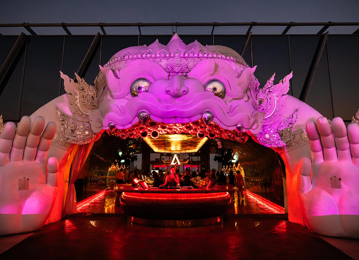 Large white sculptural entrance shaped like a mythical Thai creature glowing with pink and red lighting at a rooftop bar in Bangkok. A curved illuminated cocktail bar sits beneath the dramatic arch while guests gather inside the futuristic lounge at dusk.