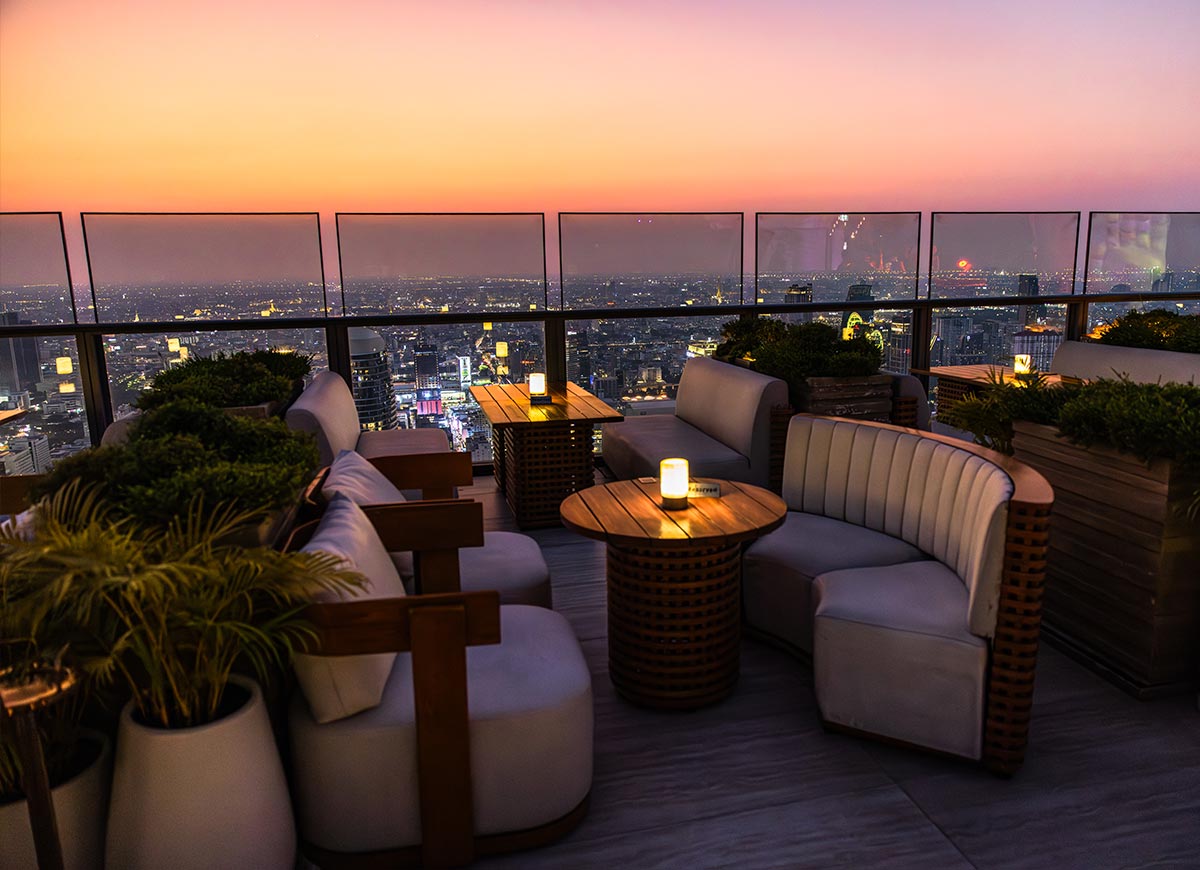 Rooftop lounge seating area with curved white chairs and candlelit tables overlooking the Bangkok skyline at sunset. Warm pink and orange skies glow behind the glass railing while potted greenery creates a cozy atmosphere above the city.