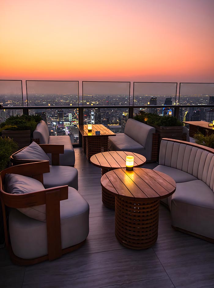 Rooftop lounge seating area with cushioned chairs and round wooden tables, each lit by a small candle, overlooking a sprawling city skyline at sunset. A glass railing frames the view as the sky fades from warm orange to soft pink. The setting feels intimate and relaxed, designed for quiet drinks above the city.
