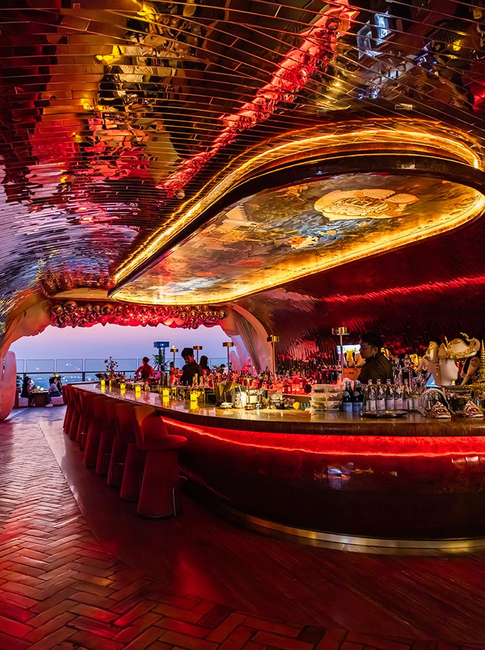 Sleek rooftop bar with a curved counter glowing in red and gold lighting, lined with plush stools and filled with bottles and glassware. A reflective ceiling and illuminated artwork overhead create a dramatic, immersive atmosphere. Guests gather around the bar while city views are visible at the open edge of the space.