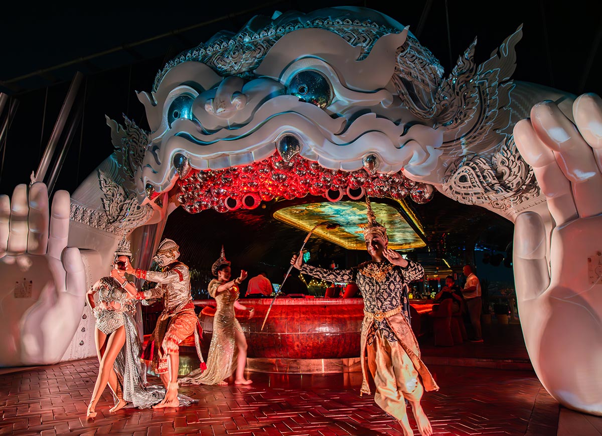 Performers in ornate traditional costumes dance beneath a large sculptural mask entrance with glowing red lights inside its open mouth. A circular bar glows in the background while oversized hands frame the dramatic scene on either side. The setting feels theatrical and immersive, blending cultural performance with a vibrant nightlife atmosphere.