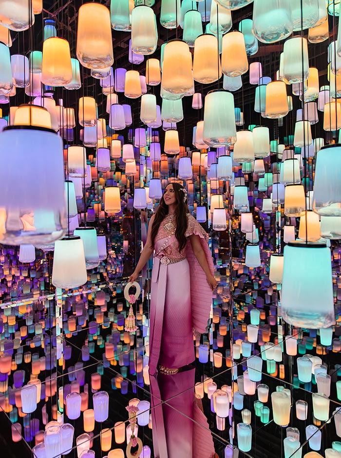 Mirrored room filled with hanging lantern lights in soft pastel colors, creating endless reflections across the floor and walls. A woman in a pink dress stands among the lights, holding a small accessory and looking upward. The immersive space feels dreamlike and vibrant, blending art, light, and reflection.