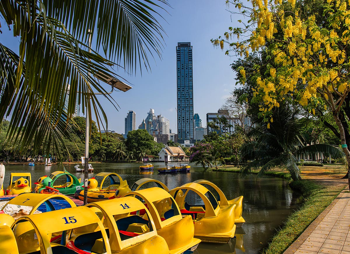 Park lake with bright yellow duck shaped pedal boats docked along the edge, surrounded by palm trees and flowering greenery. A small pavilion sits across the water with modern city skyscrapers rising in the background. The scene blends a peaceful urban park setting with playful recreational boats on the lake.