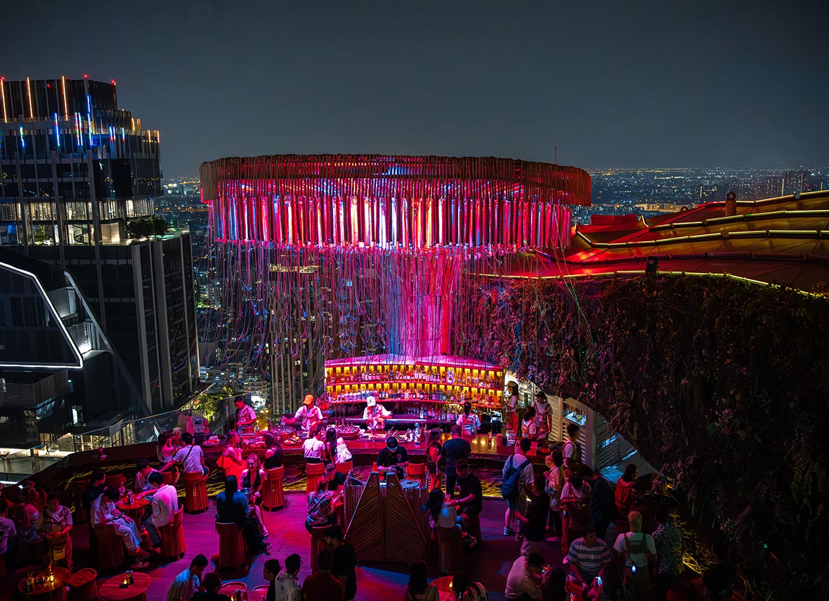 Vibrant rooftop nightclub in Bangkok glowing with pink and purple neon lighting around a circular hanging installation above the central bar. Crowds gather around cocktail tables while the illuminated city skyline stretches into the distance at night.