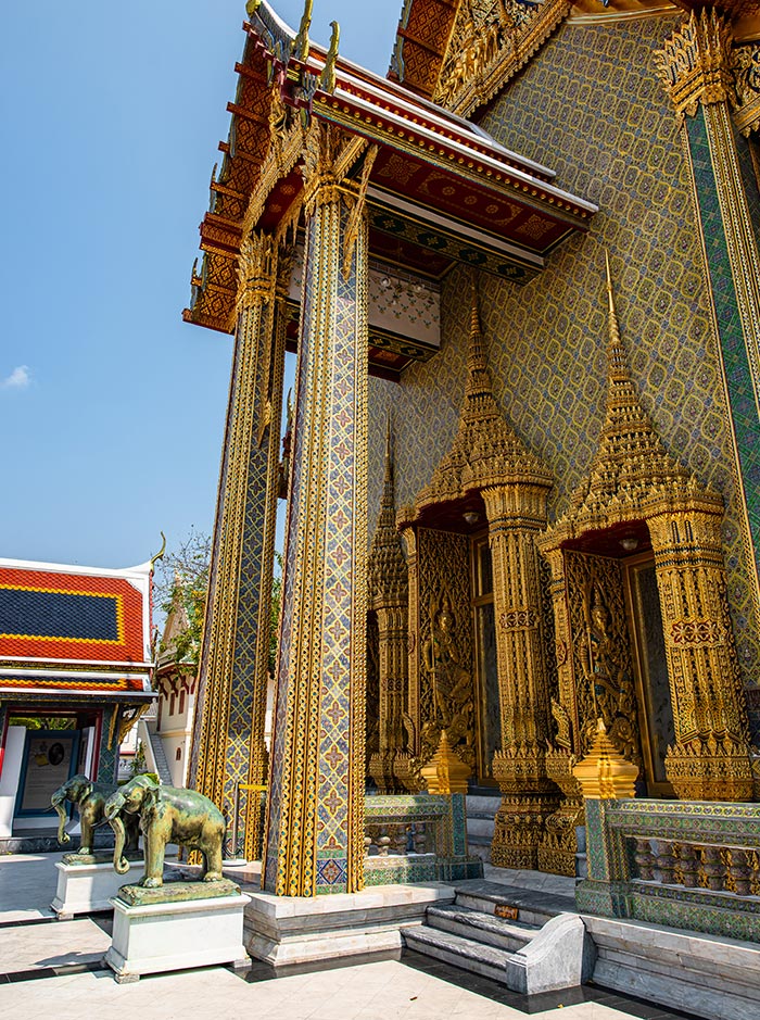 Ornate Thai temple entrance with intricate gold carvings and patterned columns stands beside a pair of small elephant statues at the base of the steps. Detailed spires and mosaic walls shimmer in the sunlight against a clear blue sky.