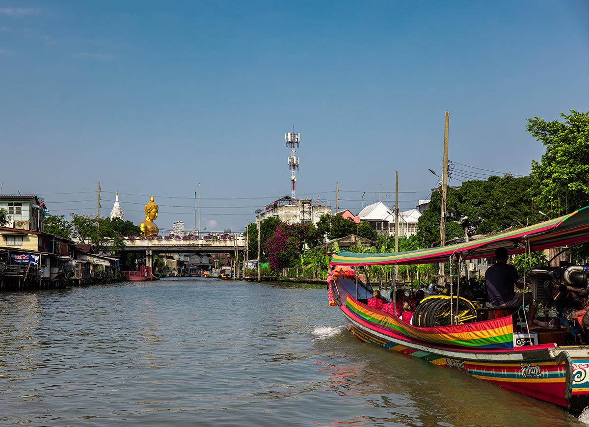 Colorful long tail boats travel along a canal lined with homes and greenery under a clear blue sky. A large golden Buddha statue rises above the waterfront in the distance, showcasing a scenic view of Bangkok's waterways and local life.