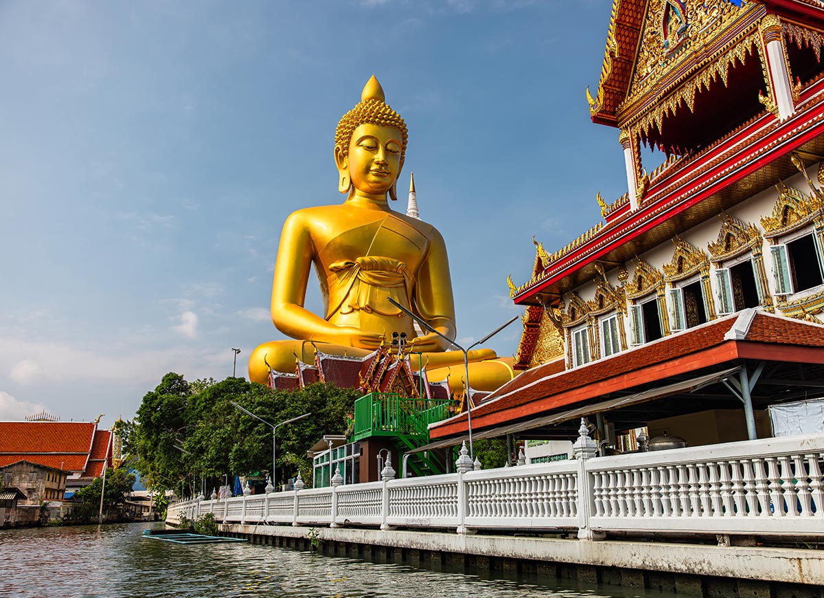 Large golden Buddha statue seated beside a riverside temple complex with ornate red and gold detailing along the building. A white fence lines the water’s edge in the foreground under a partly cloudy sky.
