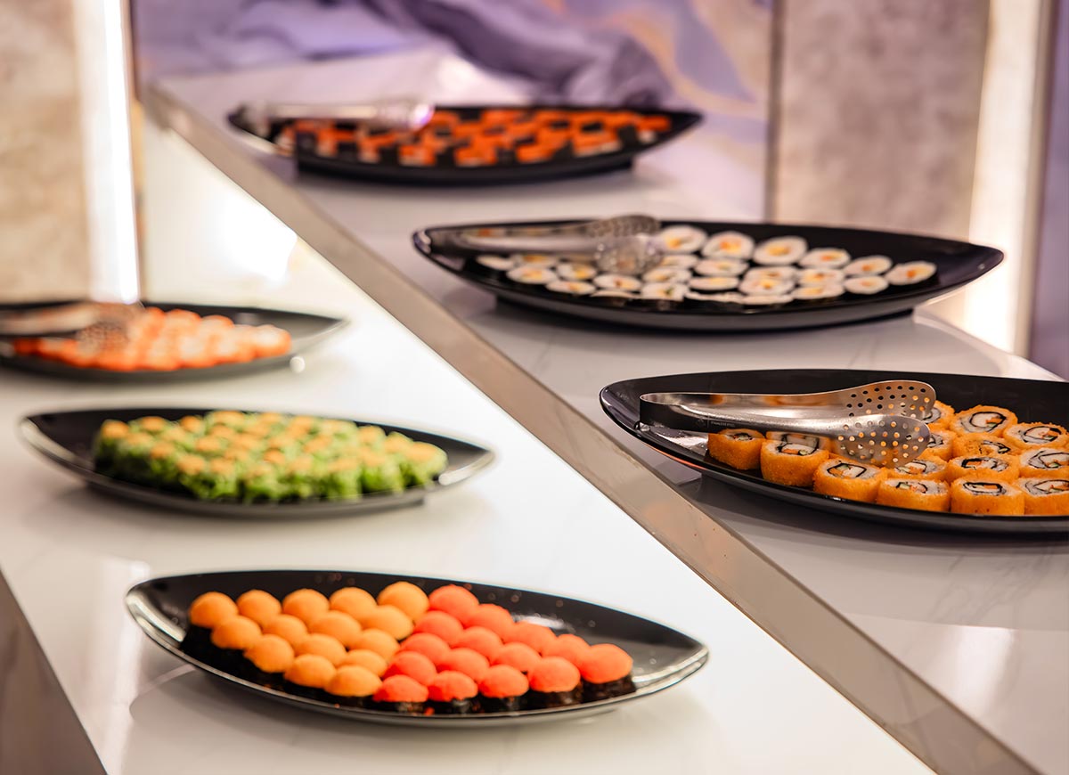 Buffet display with neatly arranged sushi rolls on black plates set along a modern white serving counter. Metal tongs rest on the trays as colorful varieties of sushi are presented in an elegant dining setting.