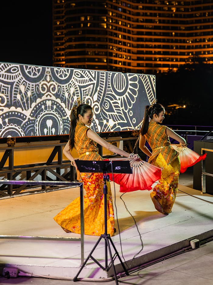 Two dancers in traditional outfits perform on a rooftop stage at night, holding red and white fans mid movement. A lit city building and patterned backdrop frame the performance under warm evening lighting.