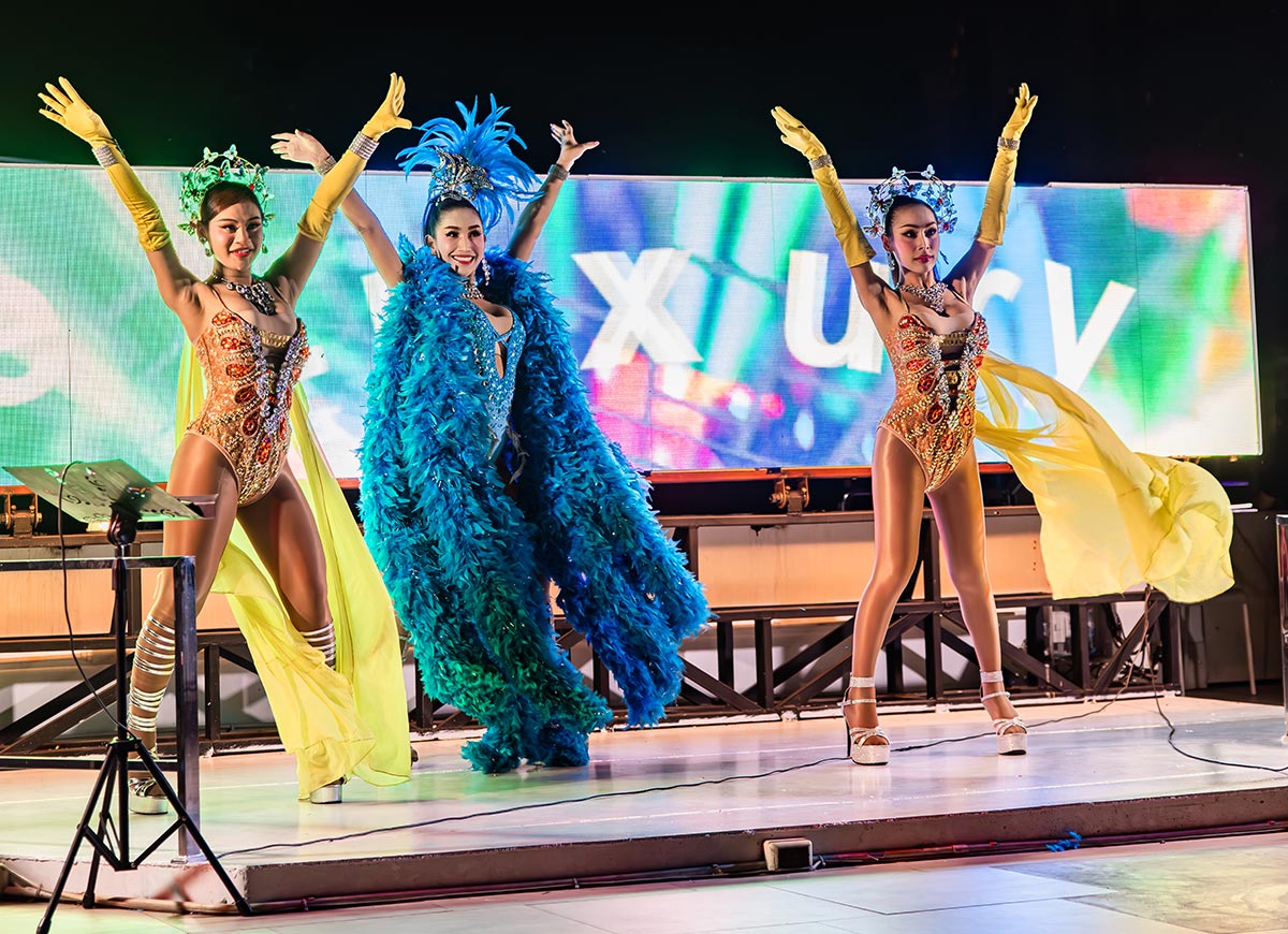 Three performers in vibrant costumes pose on a stage with arms raised, wearing embellished outfits with headpieces and flowing fabric. A colorful digital screen glows behind them, adding energy to the lively dance performance.