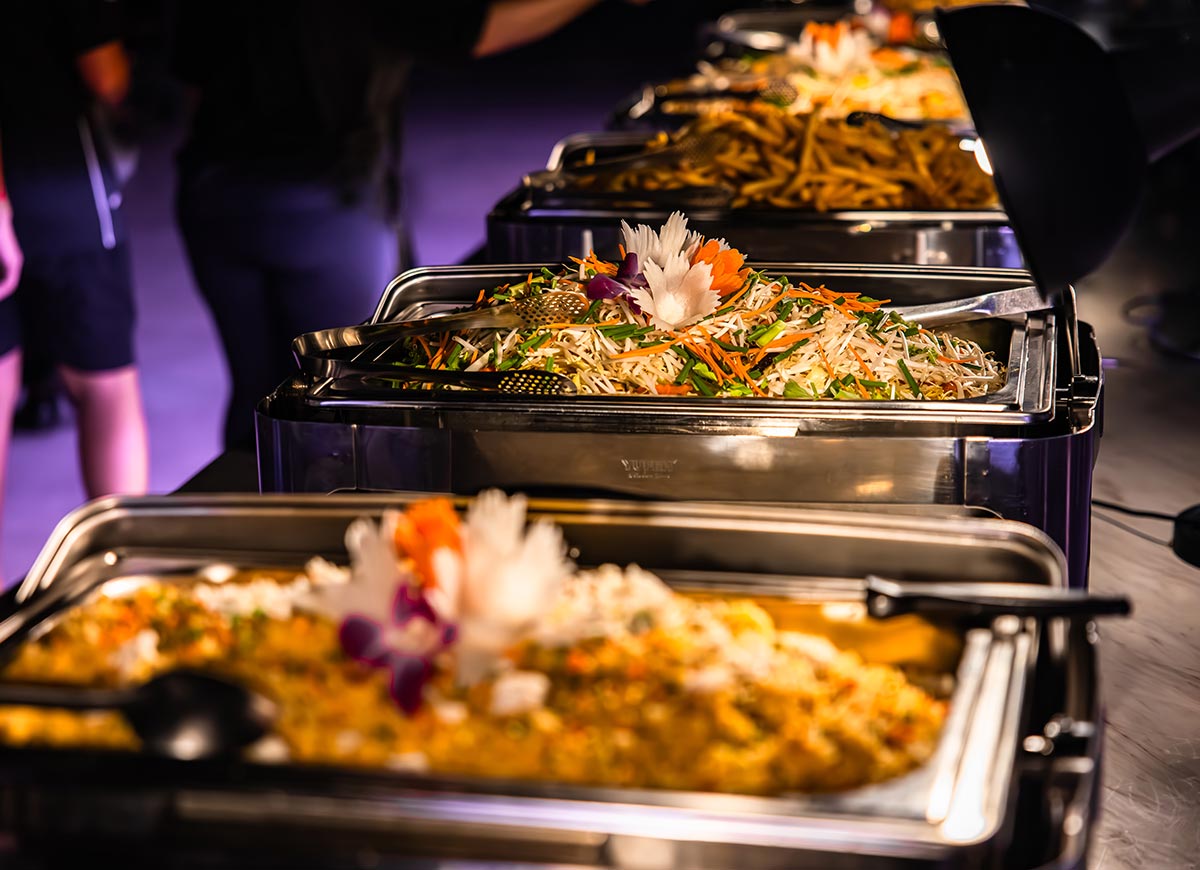 Buffet spread with stainless steel trays filled with stir fried noodles and vegetable dishes, garnished with carved flowers and herbs. Serving utensils rest on top as guests move through the line in a softly lit dining setting.
