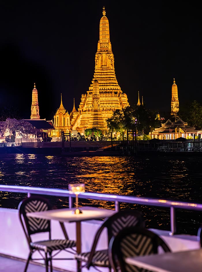 Illuminated temple complex with a tall golden spire glows against the night sky across a river. In the foreground, a candlelit table and chairs on a boat overlook the shimmering water reflecting the lights.
