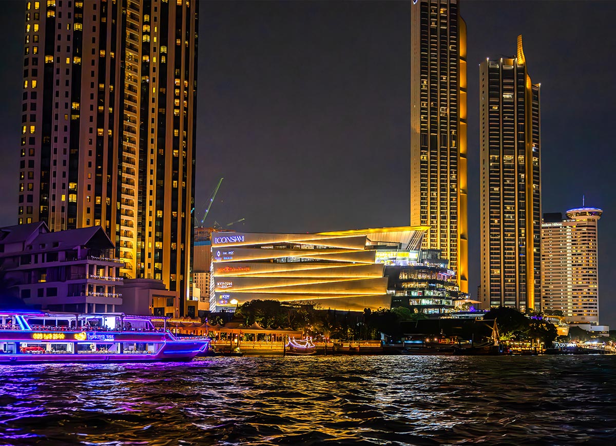 Nighttime cityscape with illuminated high-rise buildings and a brightly lit shopping complex labeled “ICONSIAM” along the waterfront. A colorful boat passes on the river in the foreground, reflecting neon lights on the water.