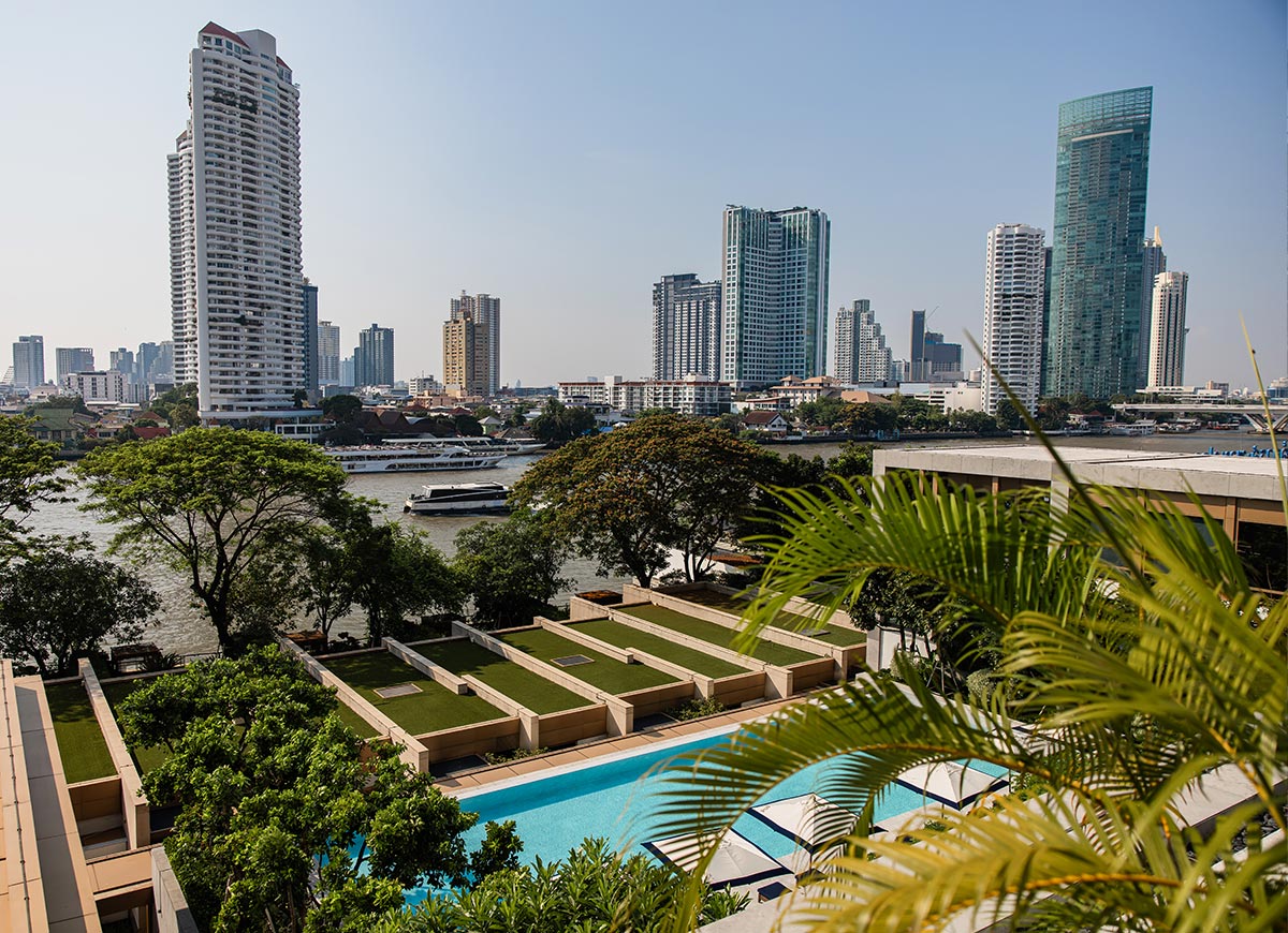 Riverside city skyline with modern high rise buildings rises beyond a tree lined waterfront, with boats moving along the river. In the foreground, a landscaped terrace with a long pool and palm trees overlooks the urban view under a clear blue sky.