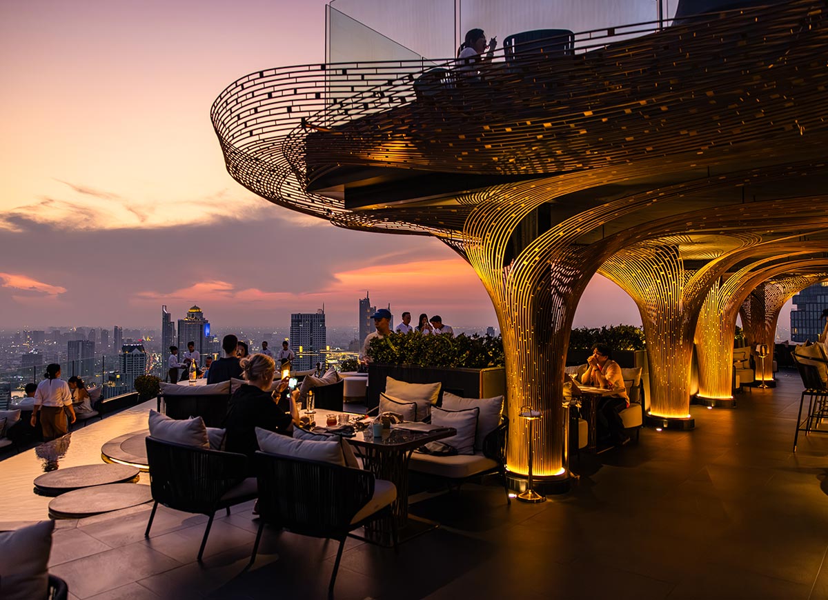 Rooftop restaurant with glowing sculptural columns overlooks a city skyline at sunset, with warm golden lighting illuminating the seating area. Guests dine and relax on cushioned chairs while the sky fades into shades of pink and orange.