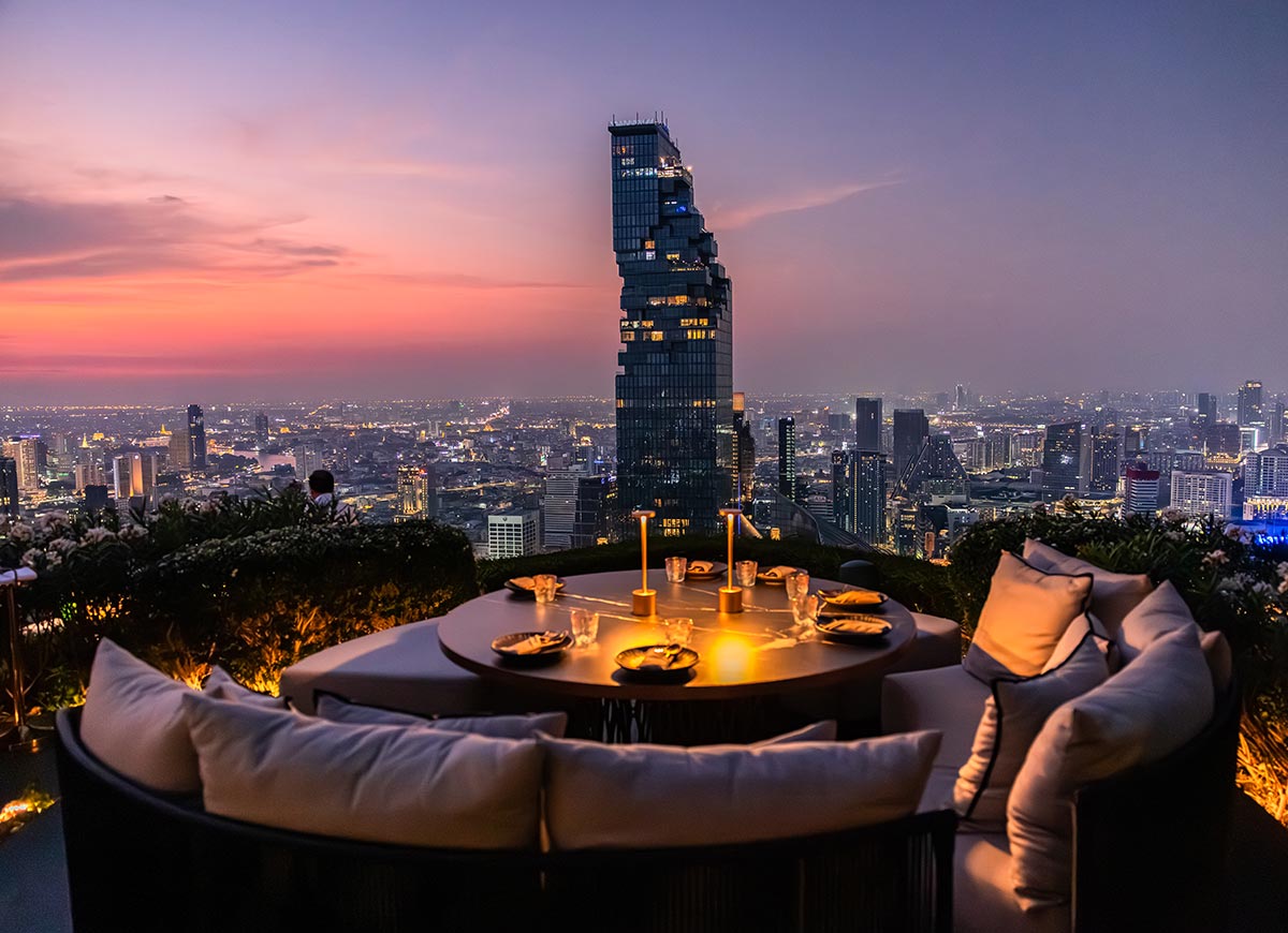 Candlelit rooftop dining table with cushioned seating overlooks a sprawling city skyline at sunset. A tall modern skyscraper rises in the center as the sky fades from pink to deep blue above the glowing city lights.