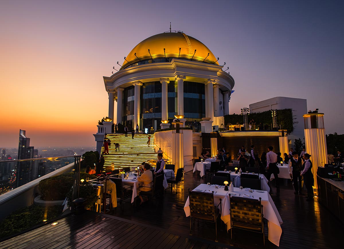 Elegant rooftop dining terrace at sunset with a grand domed building glowing in warm golden light at the top of illuminated stairs. Guests sit at white tablecloth tables while the city skyline stretches into the distance under a fading orange and purple sky.