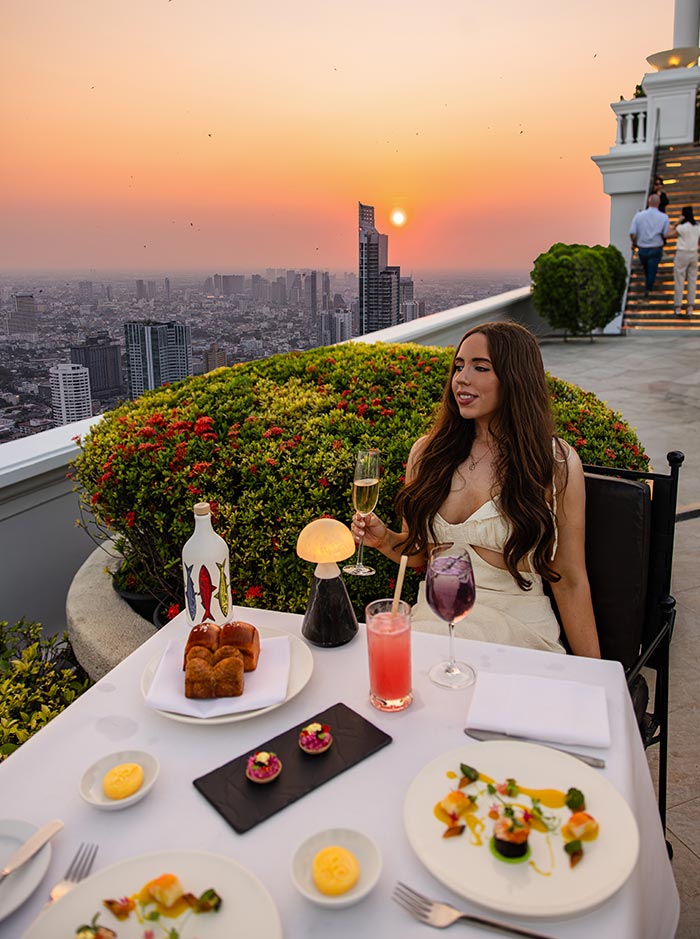 Woman in a white dress sits at a rooftop restaurant table holding a glass of wine, with plated dishes and cocktails arranged in front of her. The city skyline stretches into the distance as the sun sets in a warm orange sky behind her.