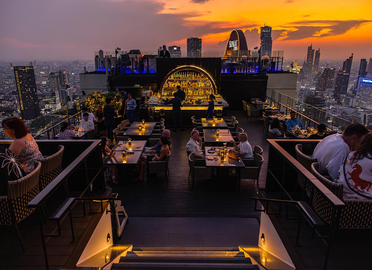 Rooftop bar at sunset with guests seated at candlelit tables surrounding a glowing central bar. The city skyline stretches into the distance under a vivid orange and purple sky as evening sets in.
