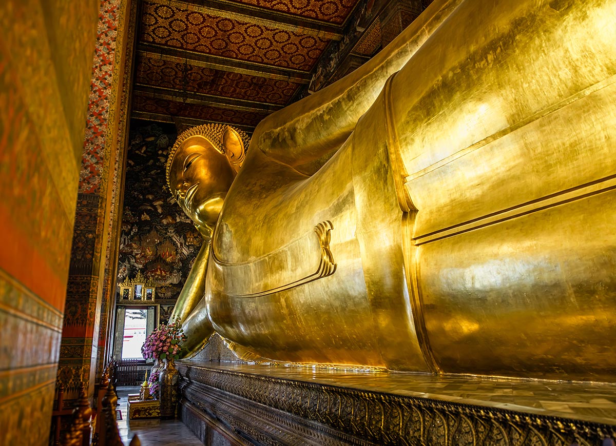 Large reclining golden Buddha statue stretches along an ornate temple interior, with its serene face resting on one hand and intricate patterns covering the surrounding walls and ceiling. Soft light reflects off the statue’s surface, emphasizing its immense size and detailed craftsmanship.