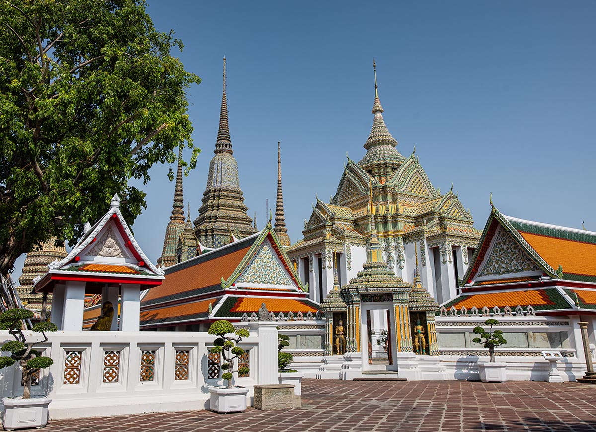 Colorful Thai temple complex with ornate rooftops and tall decorative spires rises behind a white wall and entry gate. Manicured trees and a tiled courtyard lead toward the richly detailed buildings under a clear blue sky.
