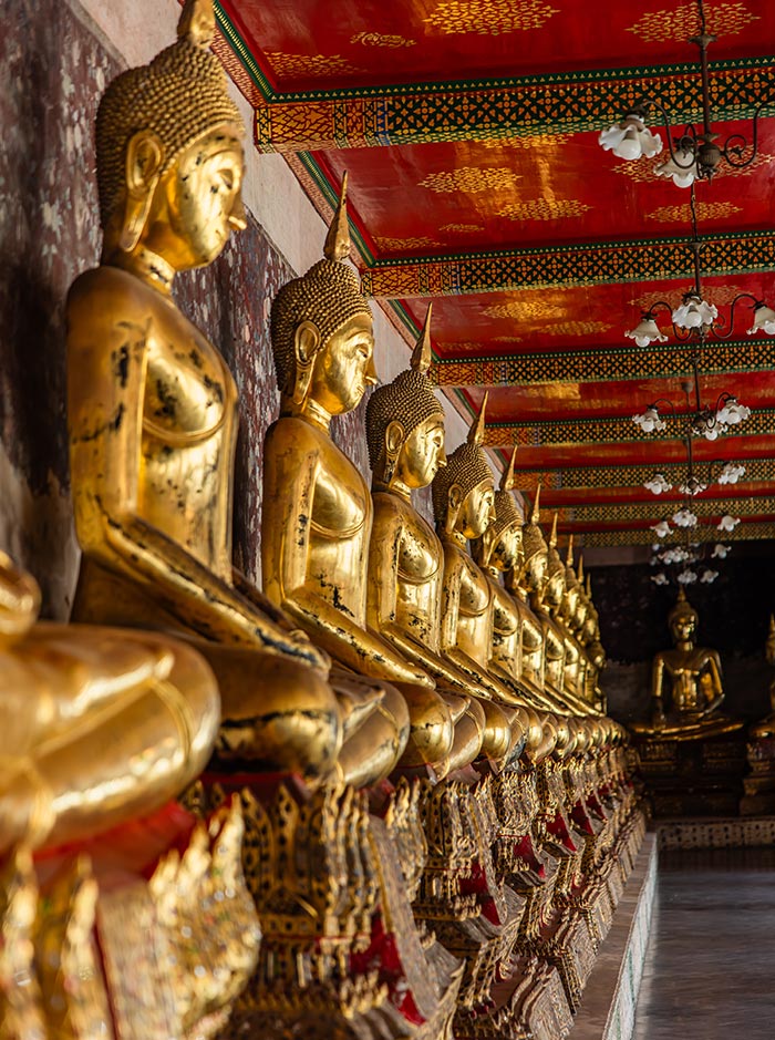 Row of golden Buddha statues seated in meditation lines a temple corridor, with intricate red and gold ceiling patterns above. The statues repeat into the distance toward a larger Buddha figure at the far end of the hall.