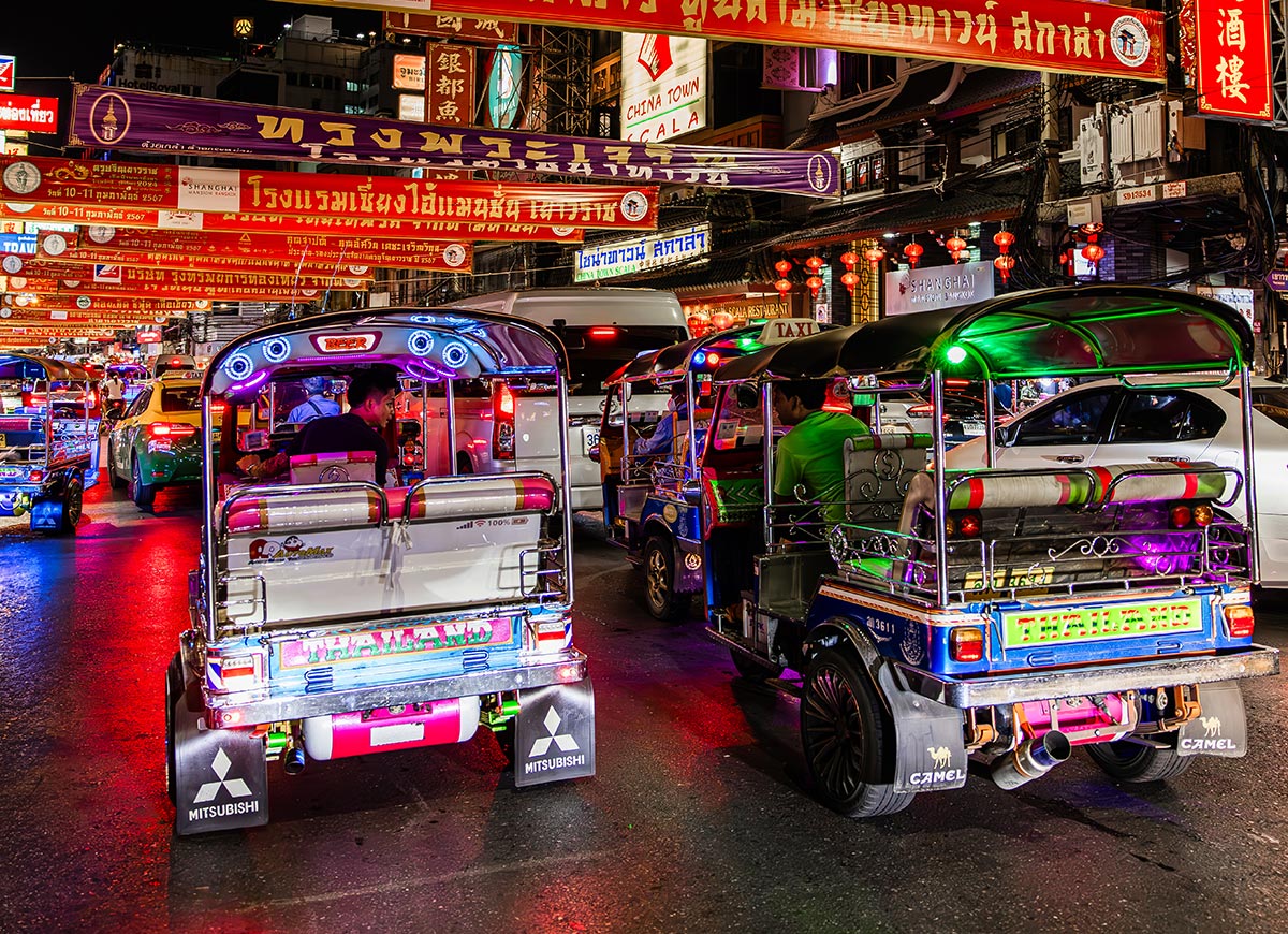 Colorful tuk tuks driving through a busy Bangkok street at night beneath rows of bright signs and banners written in Thai and Chinese. Neon lights reflect off the traffic as cars, taxis, and street restaurants fill the lively Chinatown scene.